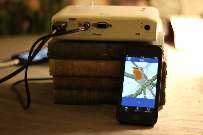 Robin sings on a mobile phone which is lent against stacks of books and a projector